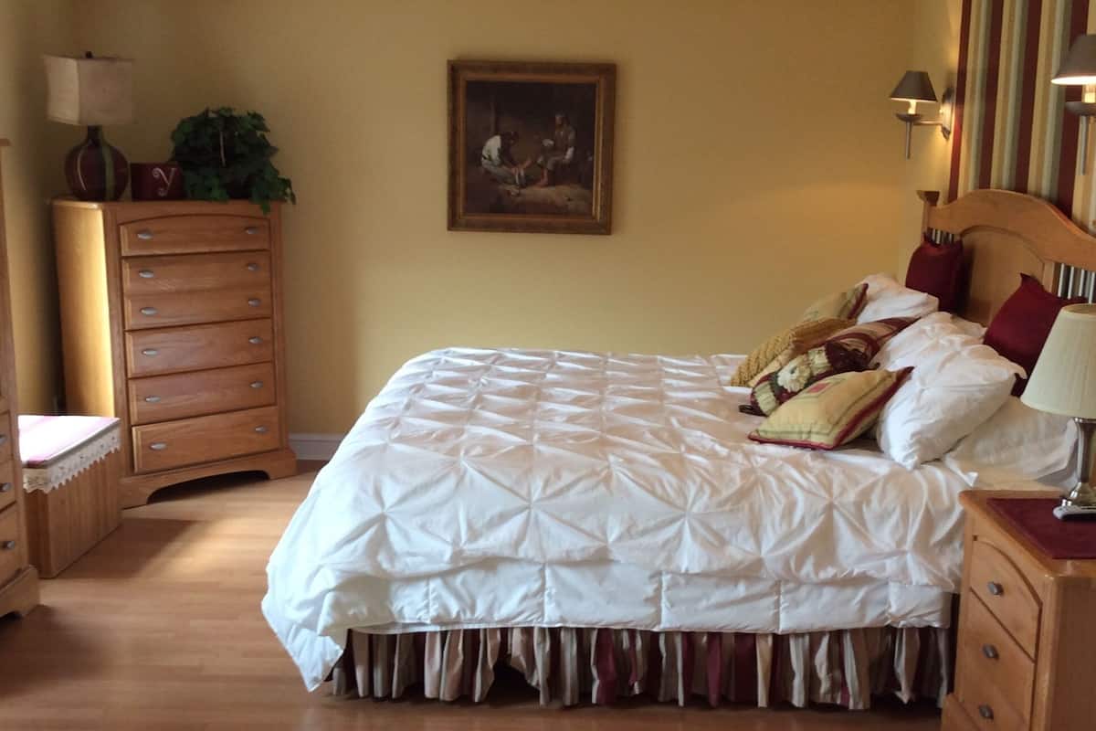 A neatly arranged bedroom with a large bed, a wooden dresser, a nightstand, and a painting on the wall.