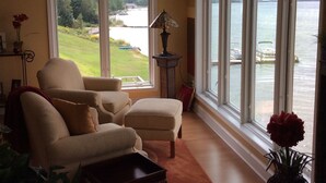 A living room with a large window overlooking a lake and greenery. There is a chair, a table, and a lamp. - Pictured Rocks Bed And Breakfast (Munising)