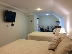 A hotel room with two beds, a flat-screen TV mounted on the wall, a desk with a lamp, and a window with blinds.