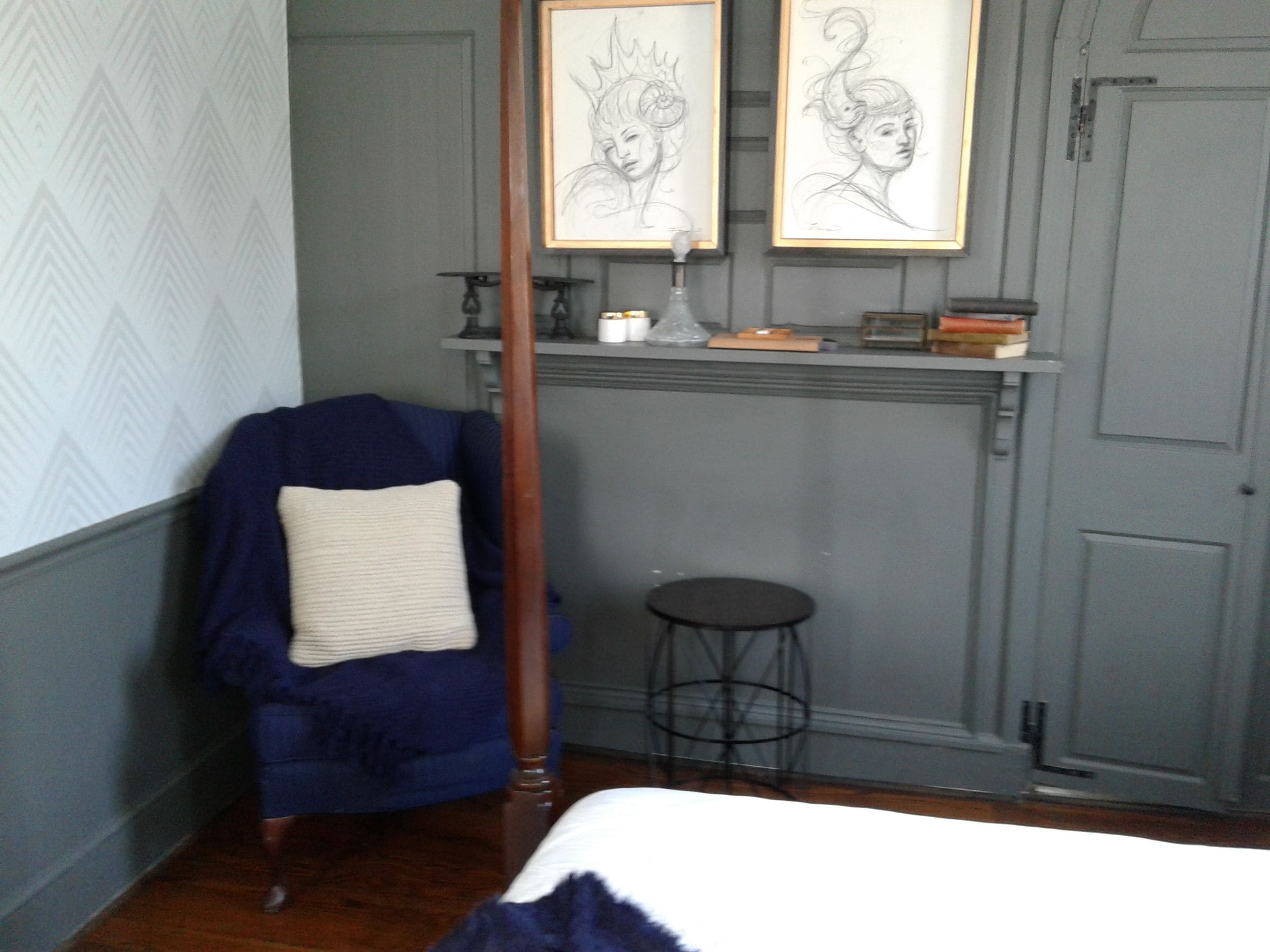 A bedroom with a blue armchair, a small stool, a bed, and two framed sketches on the wall.