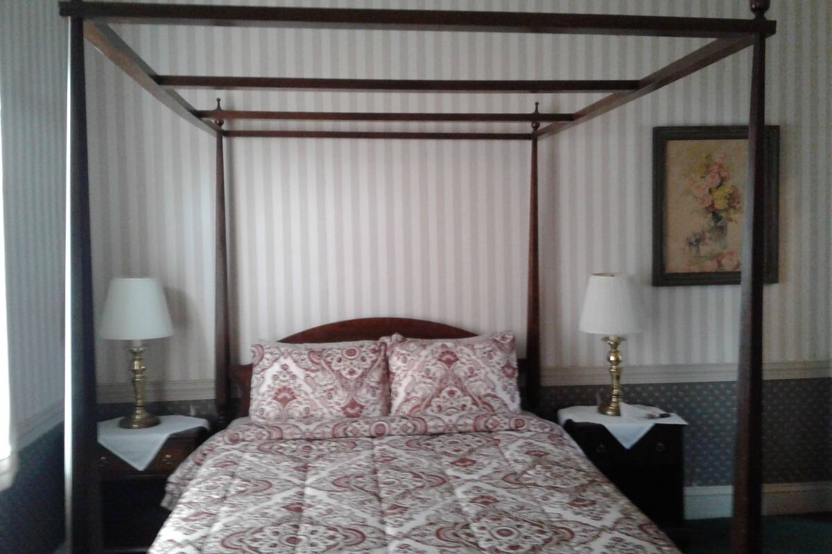 A four-poster bed with a patterned bedspread, two bedside tables with lamps, and a framed floral painting on the wall.