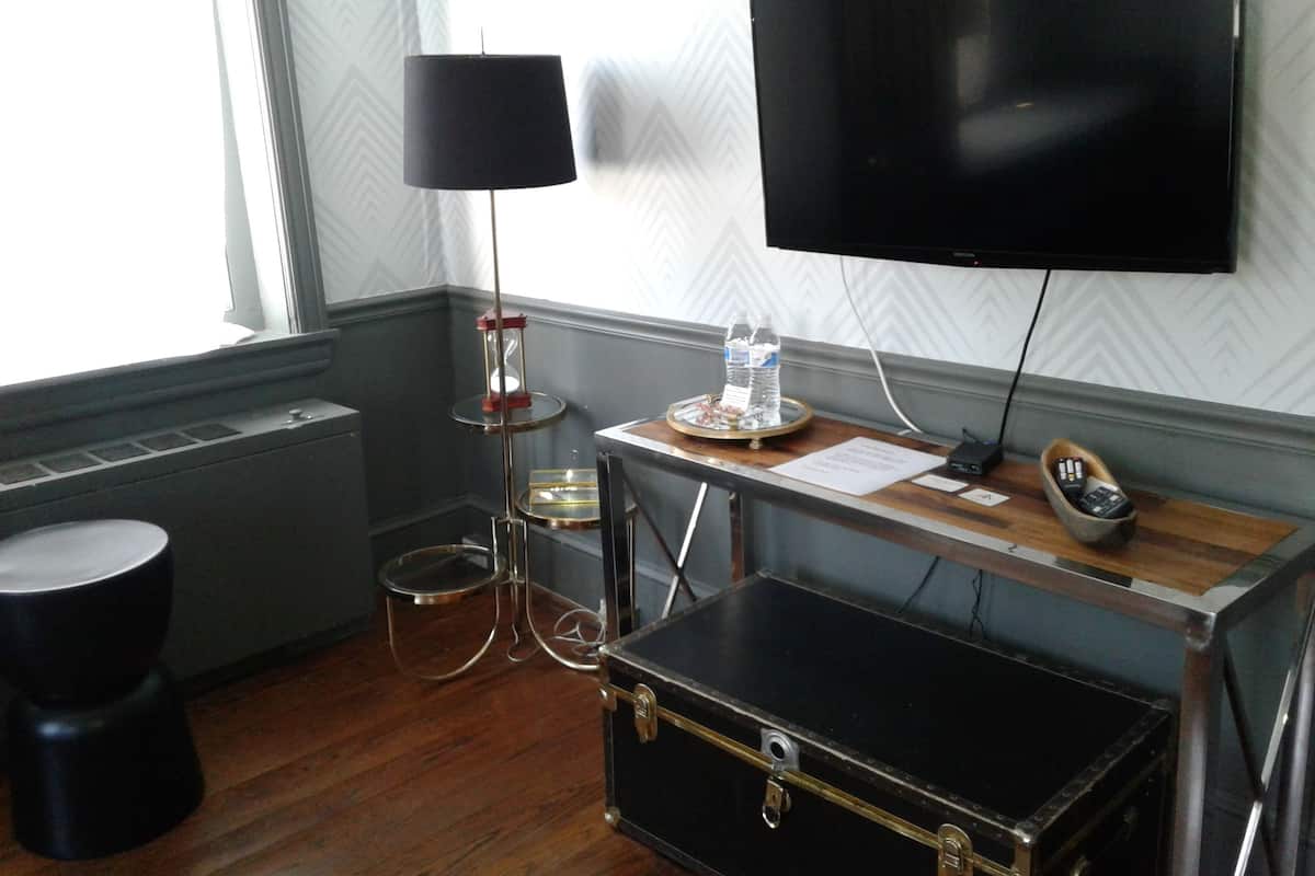 A hotel room with a wooden floor, a black suitcase, a desk with a flat-screen TV, a lamp, and a small table with a bottle of water.