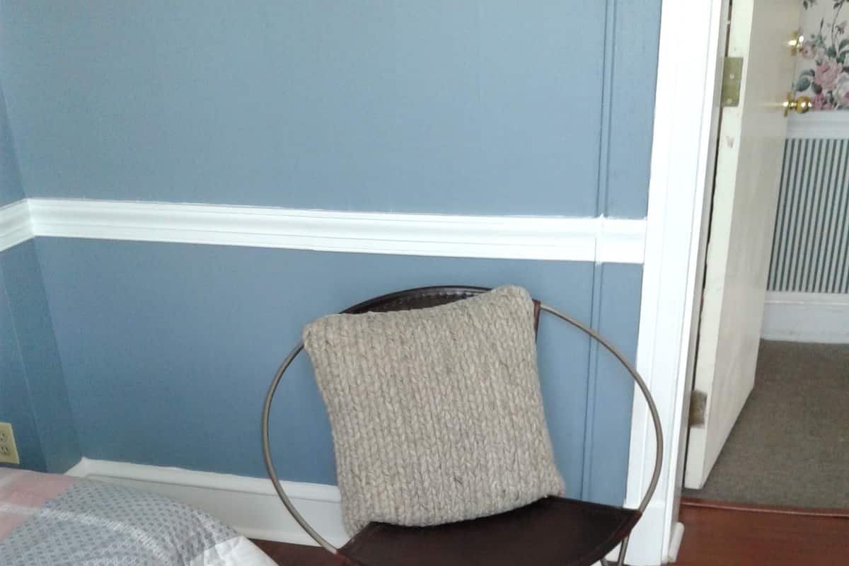 A room with a wooden floor, a bed with a white bedspread, and a chair with a grey cushion.