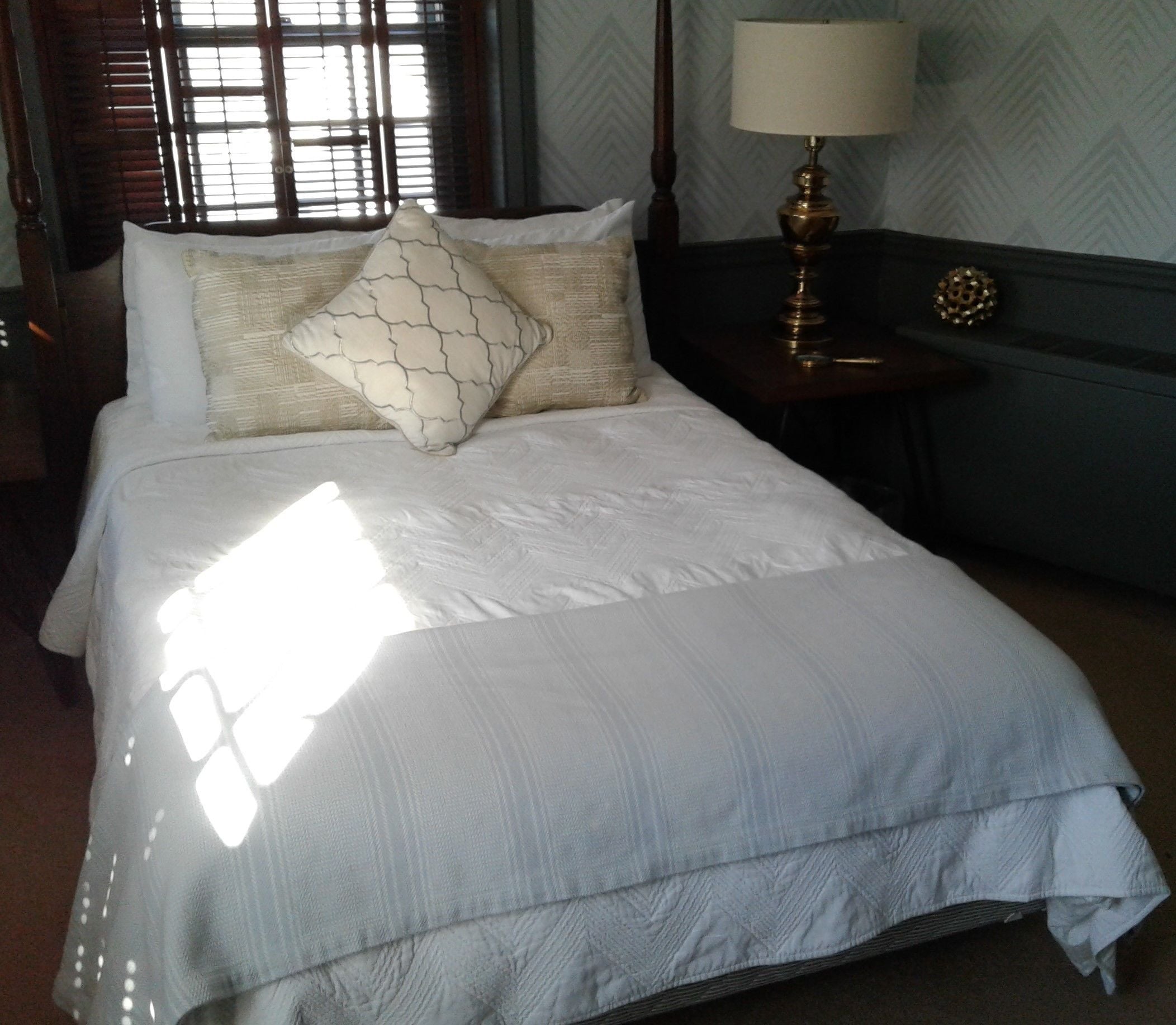 A neatly made bed with a white comforter and two pillows, a bedside table with a lamp, and a window with blinds.