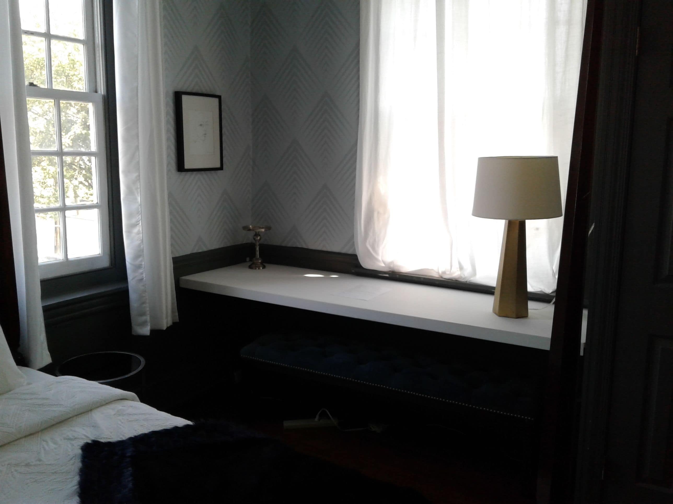 A bedroom with a window seat, a bedside table with a lamp, and a framed picture on the wall.