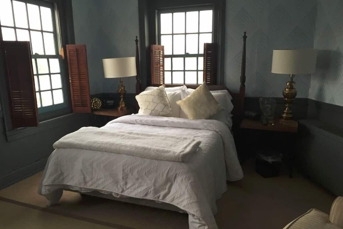 A bedroom with a large bed, two bedside tables with lamps, a window with shutters, and a chair.