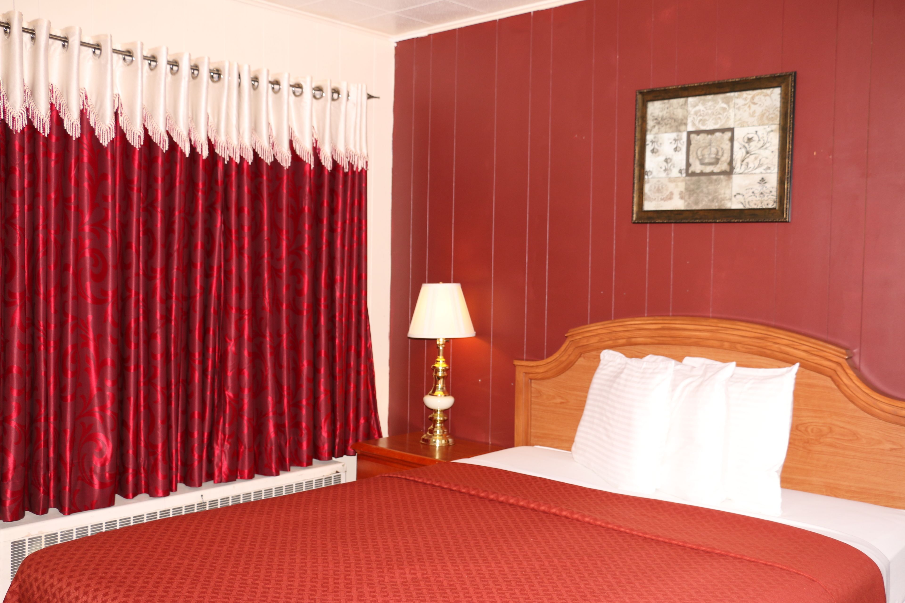 Une chambre d’hôtel avec une couvre-lit à motifs rouges, une tête de lit en bois, un oreiller blanc, un rideau rouge et un cadre photo fixé au mur.