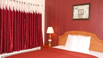 Une chambre d’hôtel avec une couvre-lit à motifs rouges, une tête de lit en bois, un oreiller blanc, un rideau rouge et un cadre photo fixé au mur.