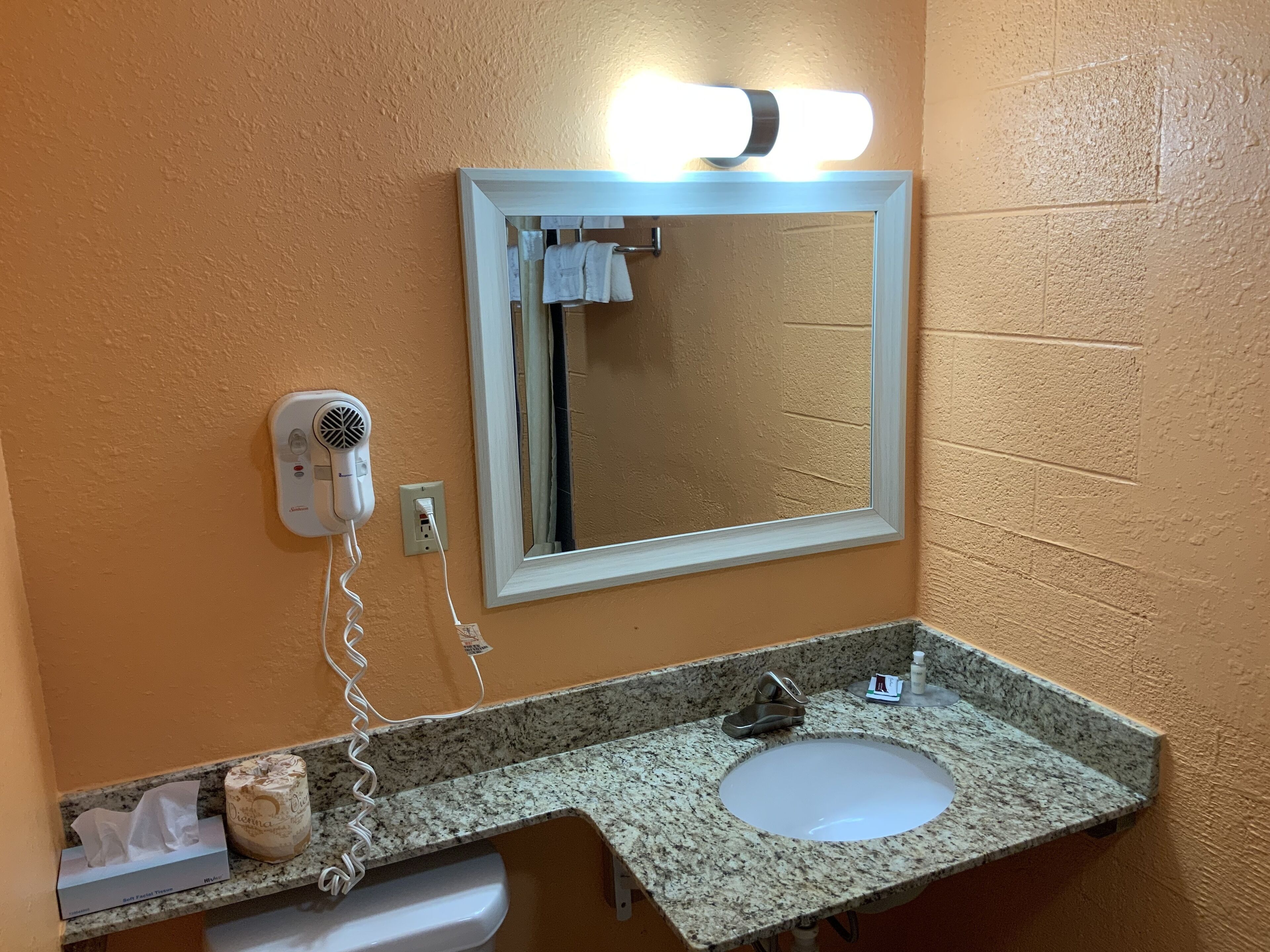Une salle de bain dotée d’un comptoir en granit, d’un lavabo blanc, d’un sèche-cheveux, d’un miroir avec éclairage intégré et d’un porte-serviettes.