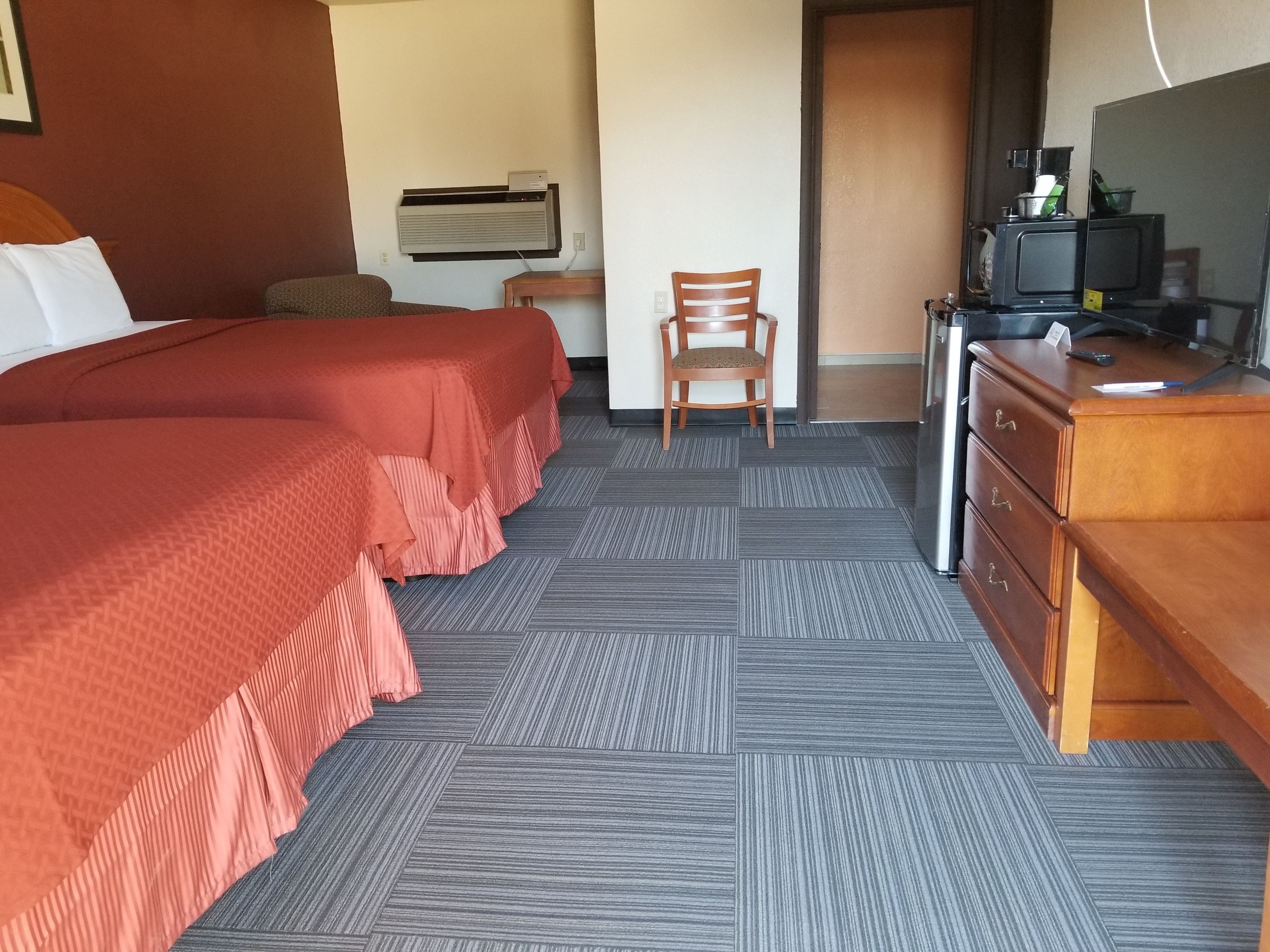 A hotel room with two beds, a wooden dresser, a chair, a desk with a computer, and an air conditioning unit on the wall.
