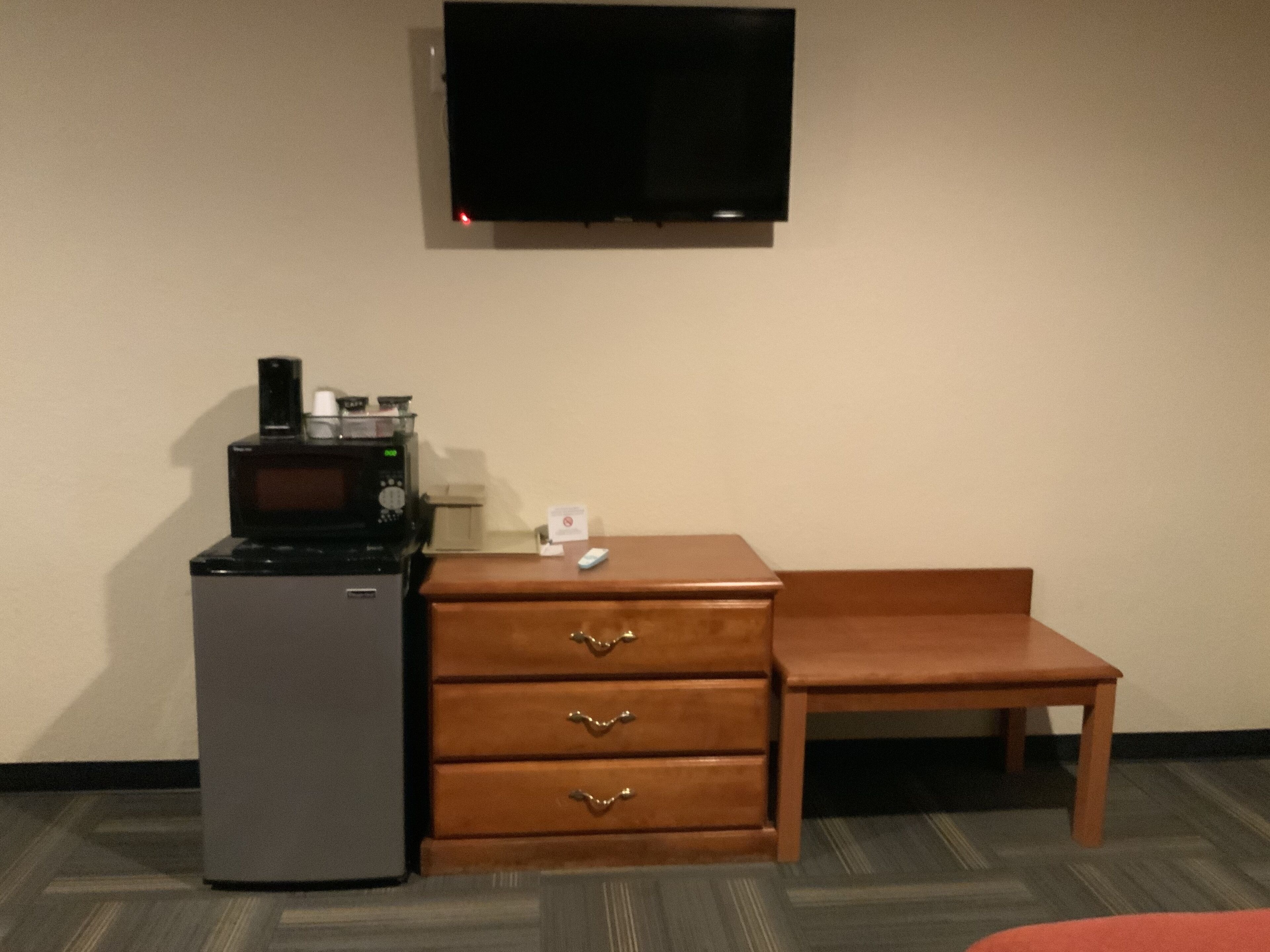 Habitación de hotel con televisor de pantalla plana en la pared, refrigerador pequeño con microondas, cómoda de madera con cajones y una silla de madera.