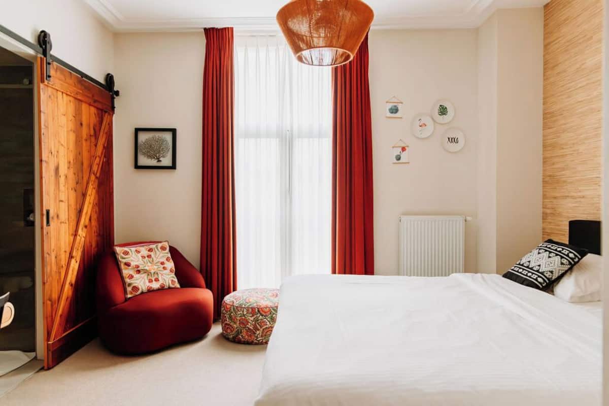 Une chambre à coucher avec un lit, un fauteuil rouge, un petit pouf, une armoire en bois et une grande fenêtre avec des rideaux rouges.