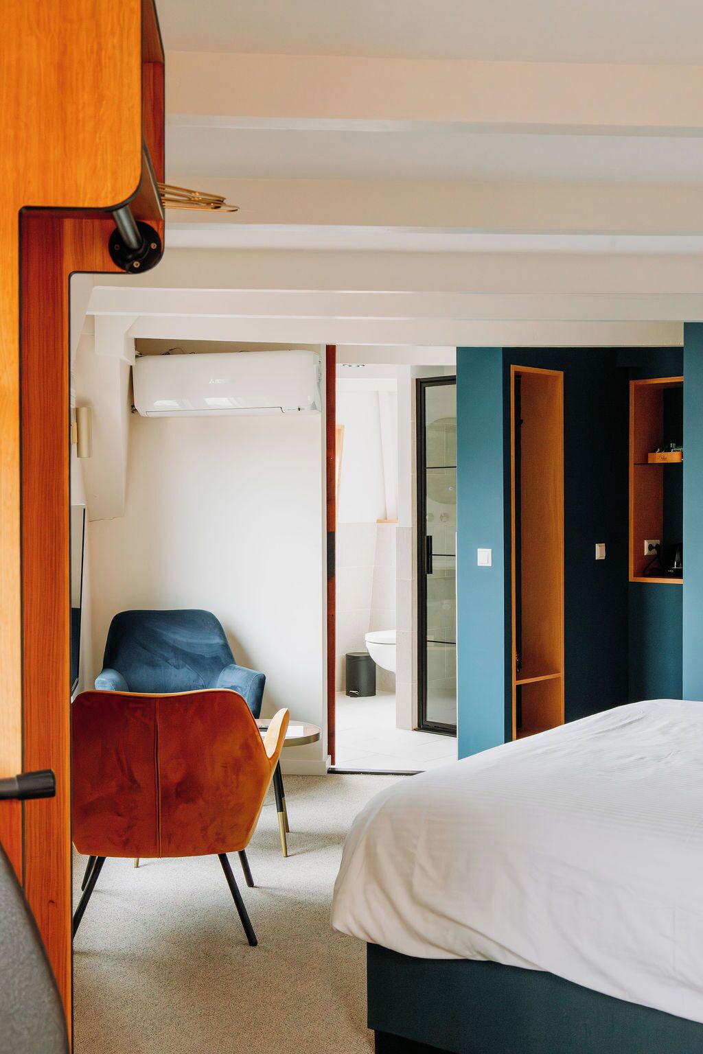 Quarto de hotel moderno com uma cama, uma poltrona azul, um guarda-roupa de madeira e um banheiro visível por uma porta aberta.