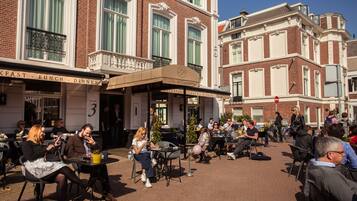 Un área de café al aire libre con mesas y sillas, un letrero de un hotel, y gente comiendo y conversando.