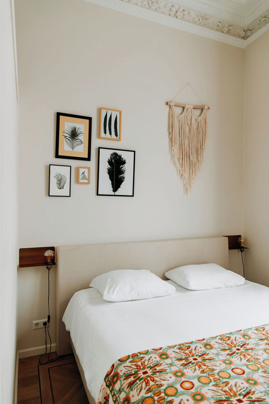 A bedroom with a bed, white pillows, a patterned blanket, and wall art including feather prints and a macrame piece.