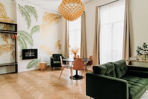 Un salon moderne avec un canapé vert, une table de salle à manger ronde entourée de chaises, un luminaire suspendu et un mur orné d’un motif de feuilles.