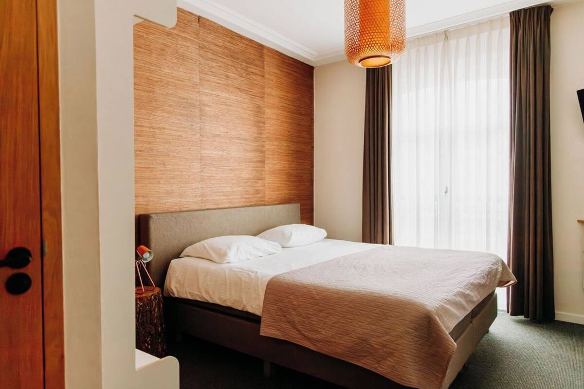 Une chambre d’hôtel avec un lit, une tête de lit en bois, un luminaire suspendu et une vue sur l’extérieur.