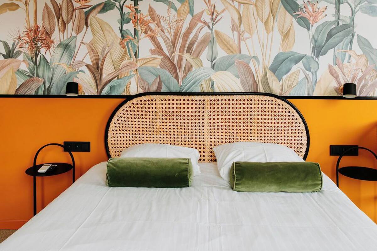 Un lit avec une literie blanche, deux oreillers verts, une tête de lit en osier et une fresque murale représentant des plantes tropicales.