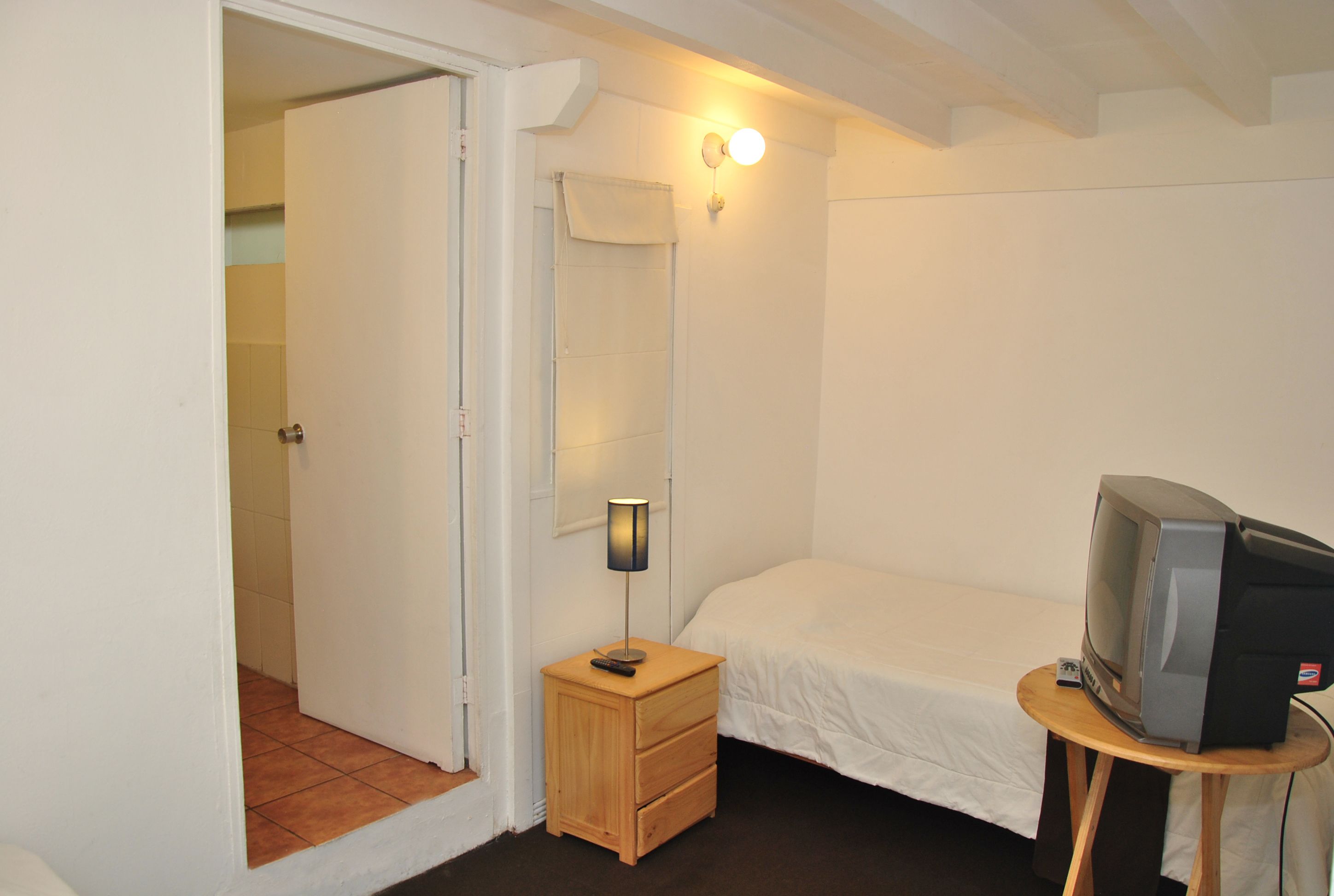 Un dormitorio con cama, una mesita de noche con lámpara, un televisor sobre una pequeña mesa y una puerta que da a otra habitación.
