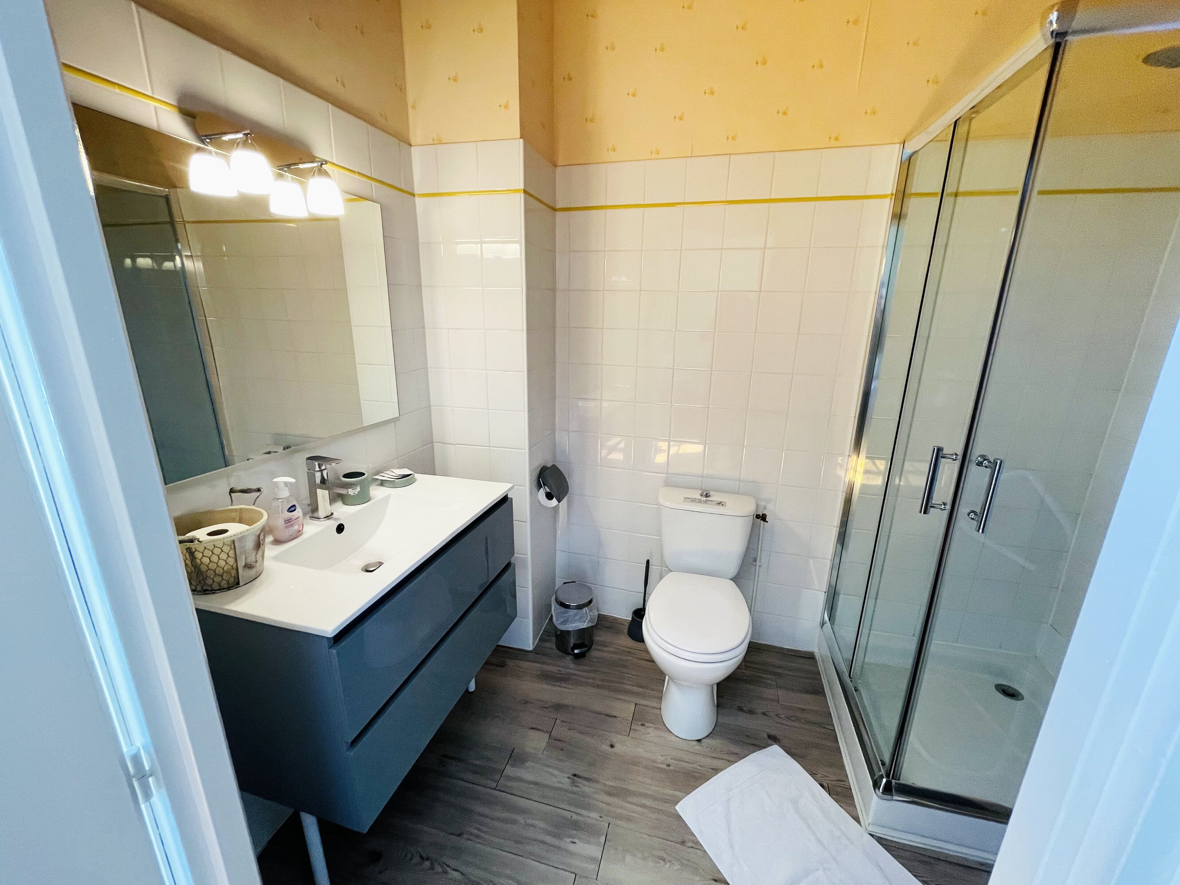 Casa de banho moderna com lavatório branco, espelho e sanita. Tem soalho de madeira e cabine de duche em vidro.