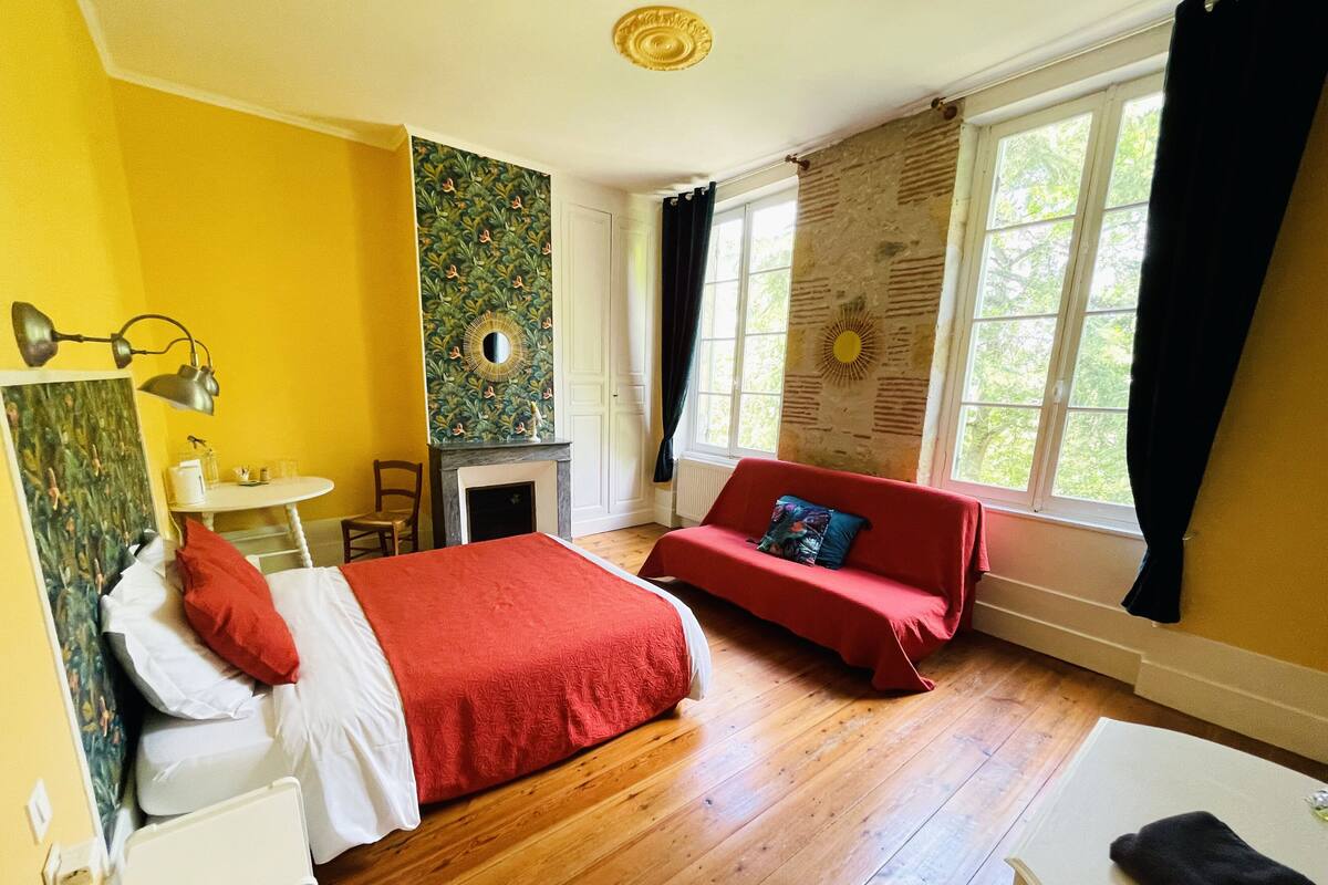 Una habitación con una cama, un sofá rojo, una mesita, un espejo y una chimenea.