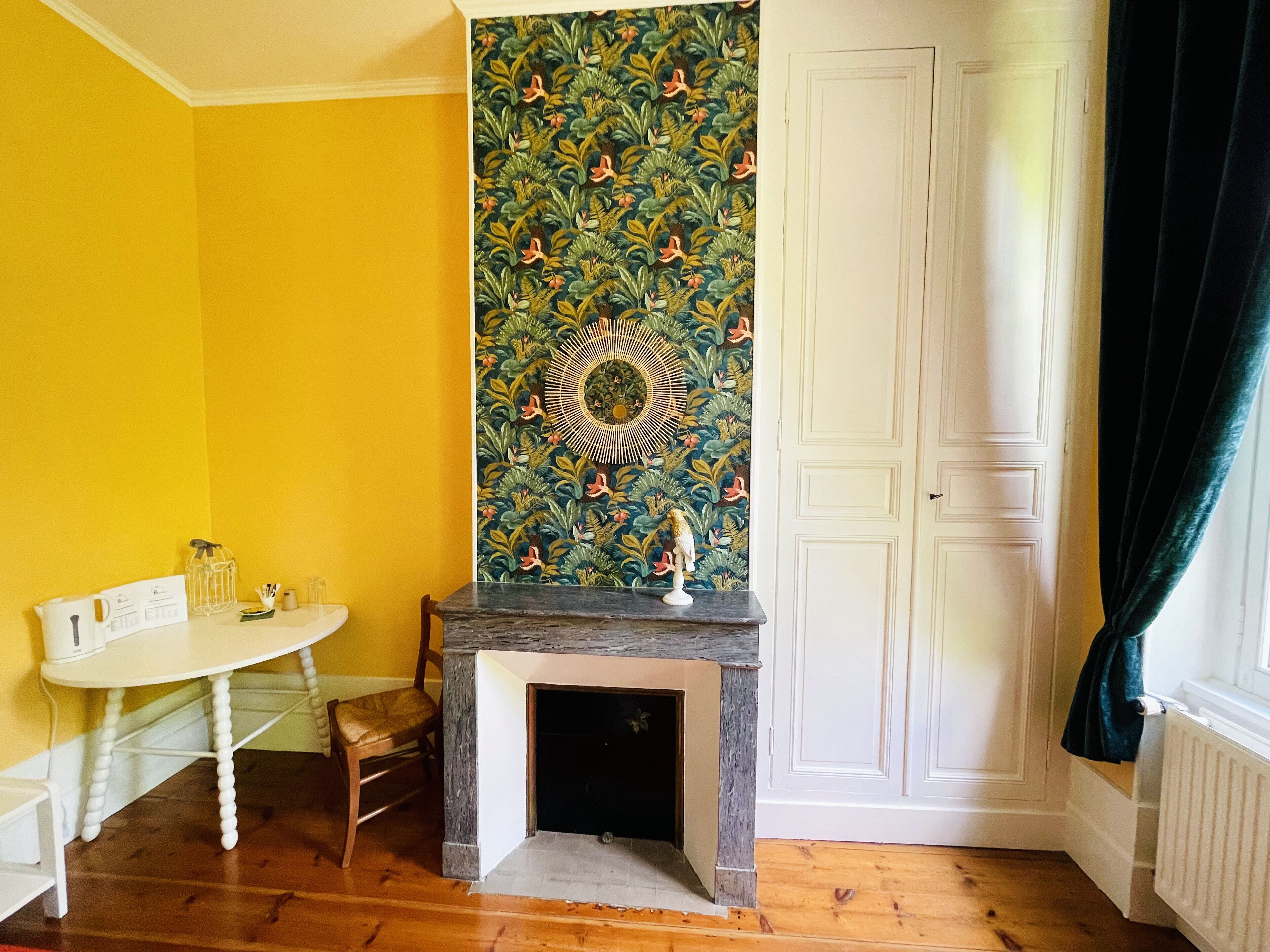Een kamer met een gele muur, een witte deur, een openhaard met decoratieve bovenkant, een kleine tafel met een stoel, en behang met een patroon.