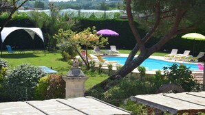 Zona de piscina con sillones de descanso y sombrillas, rodeada de vegetación y un pabellón blanco.