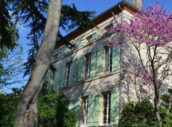 Un edificio de dos pisos con contraventanas verdes, rodeado de árboles y un árbol en flor.