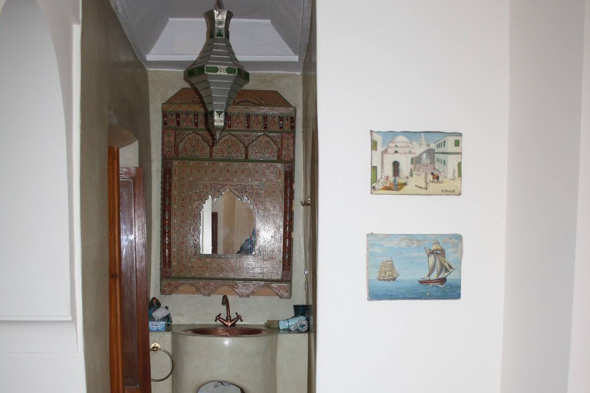 A narrow hallway with a wooden door, a small sink, a mirror, and a chair with a decorative table.