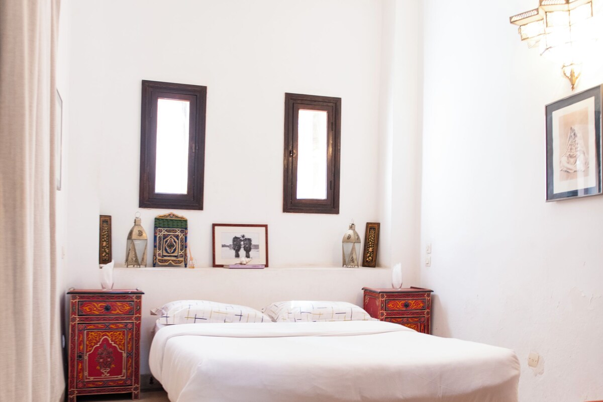 A bedroom with a bed, wooden headboard, two bedside tables, a red and gold cabinet, a patterned rug, and a wall-mounted light fixture.