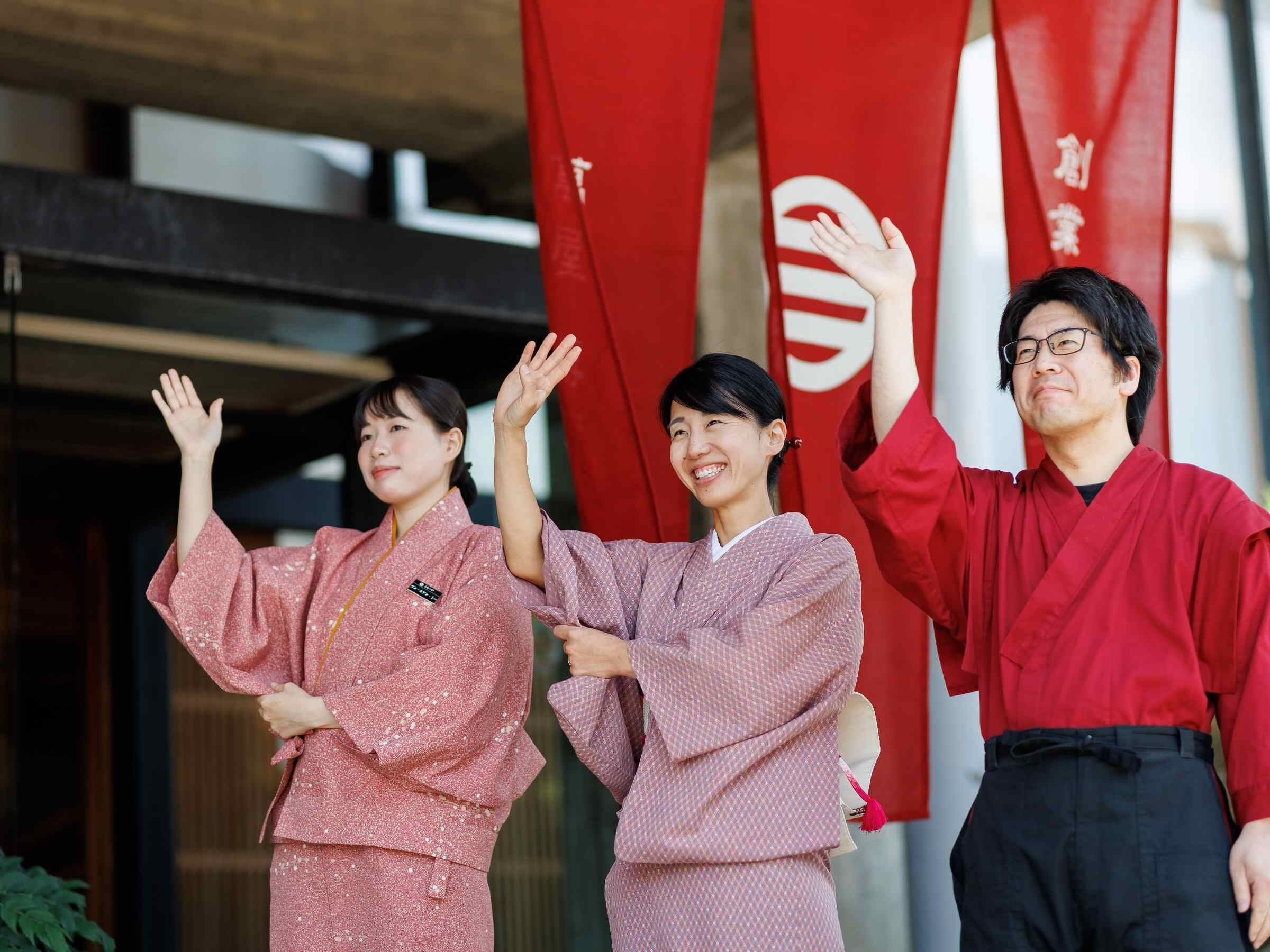 Three individuals in traditional attire standing in front of red flags with white symbols.