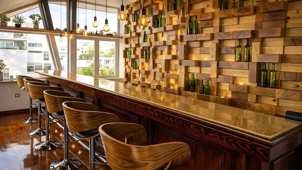 A bar area with wooden stools, a wooden wall with bottles, and a large window.