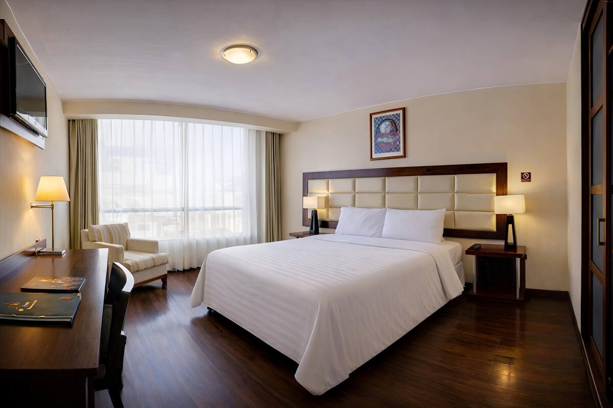 A hotel room with a large bed, a desk with a lamp, a chair, a TV, and a window with curtains.