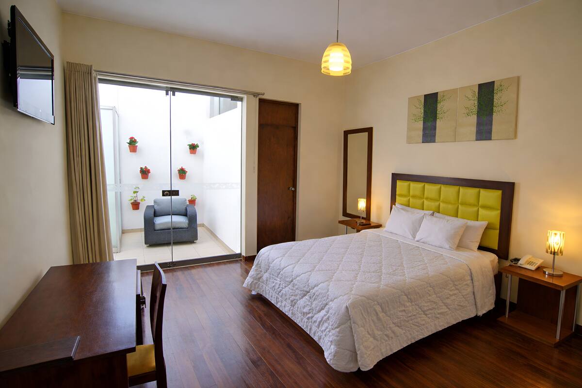 A hotel room with a bed, a desk, a chair, a TV, a bedside table, a lamp, and a balcony with potted plants.
