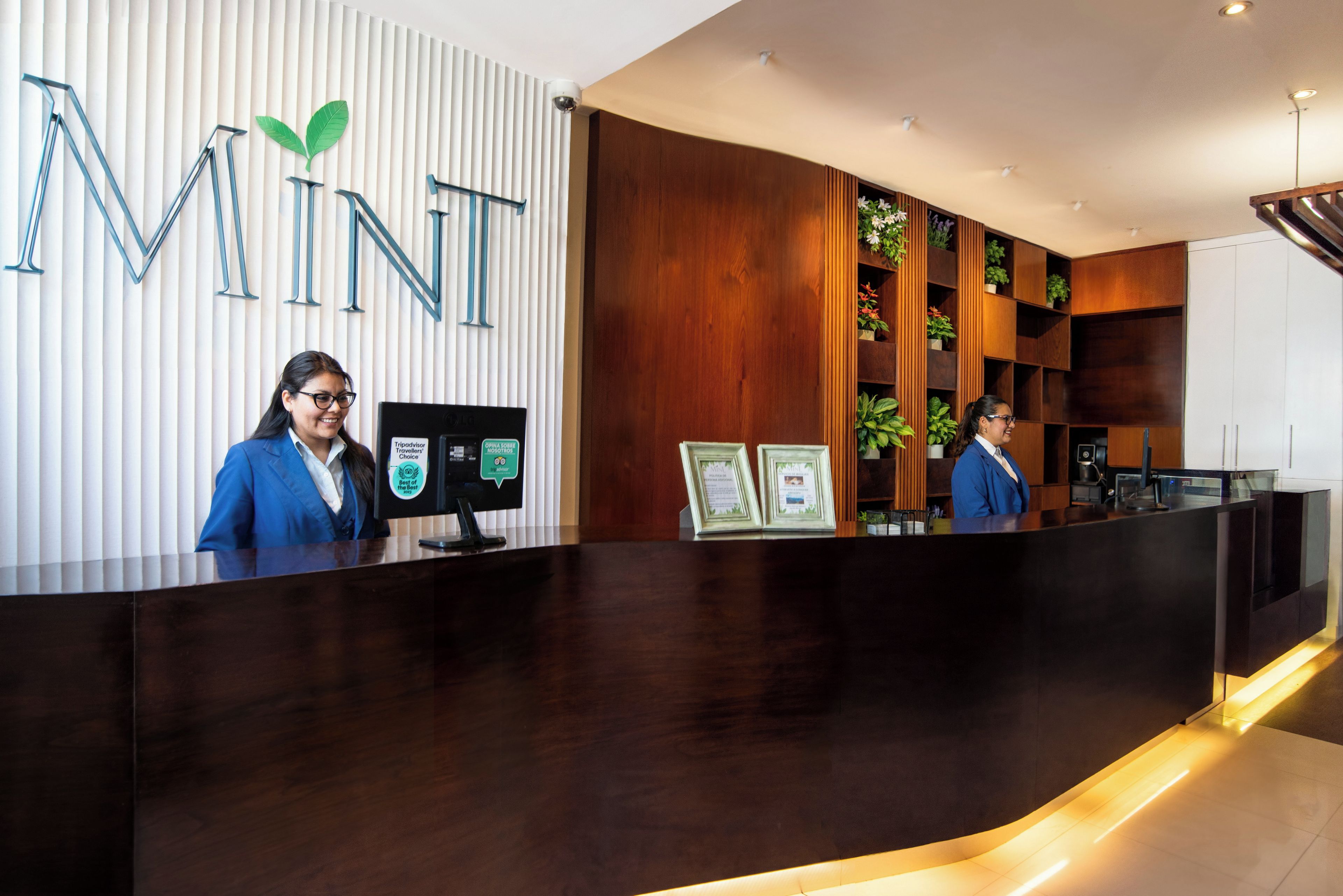 A reception desk with a wooden finish and a logo on the wall. There are two monitors and framed certificates on the desk.