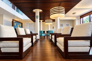 A hotel lobby with a modern design, featuring a central seating area with chairs and sofas, a wooden ceiling with slatted panels, and a view of the outdoors through large windows.