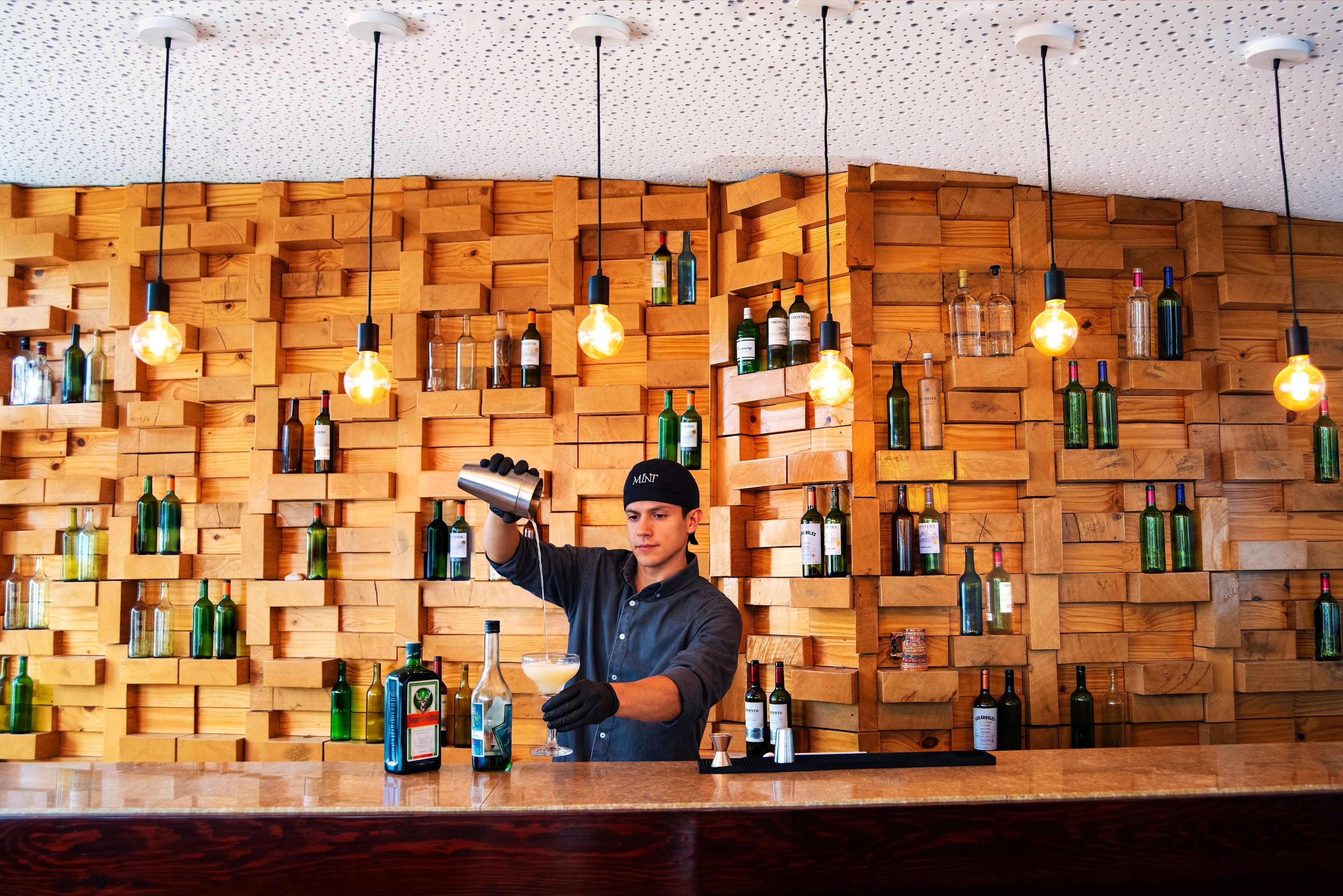 A bartender pouring a drink at a bar with wooden shelves filled with bottles.