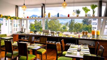 Un comedor con mesas dispuestas para el desayuno, vistas de montañas a través de amplias ventanas y varios electrodomésticos en la encimera.