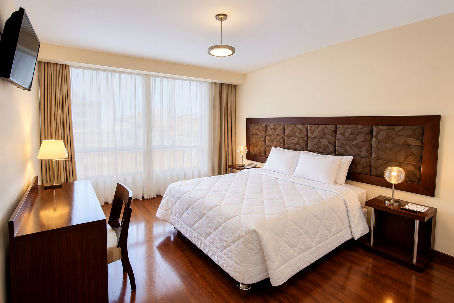 A hotel room with a large bed, a desk, a chair, a TV, and a bedside table with a lamp.