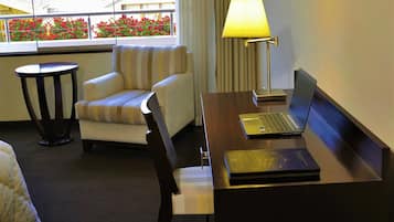 A hotel room with a bed, a desk with a laptop, a striped armchair, a lamp, and a view of buildings and flowers through the window.