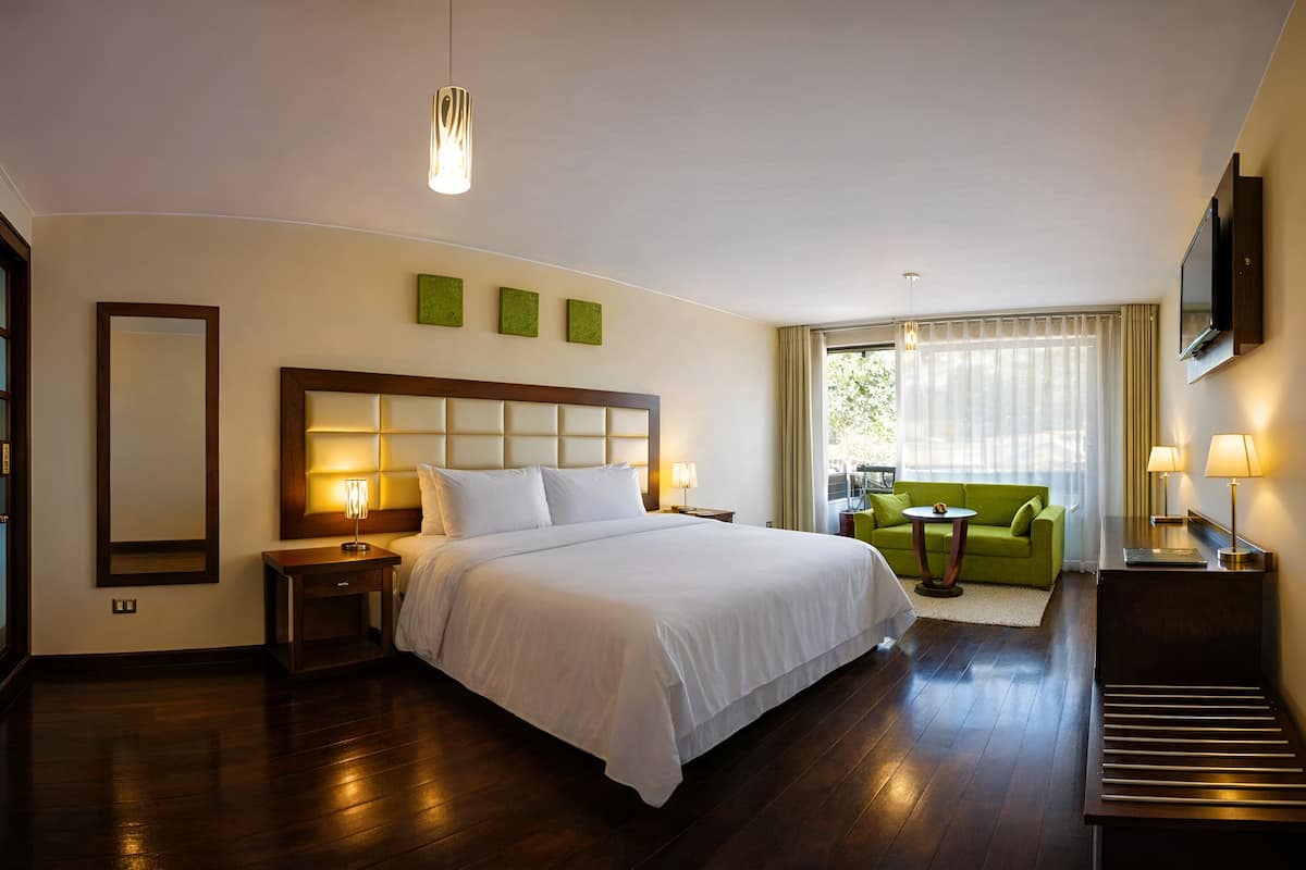 A hotel room with a large bed, bedside tables, a green sofa, a TV, and a view of the outdoors.