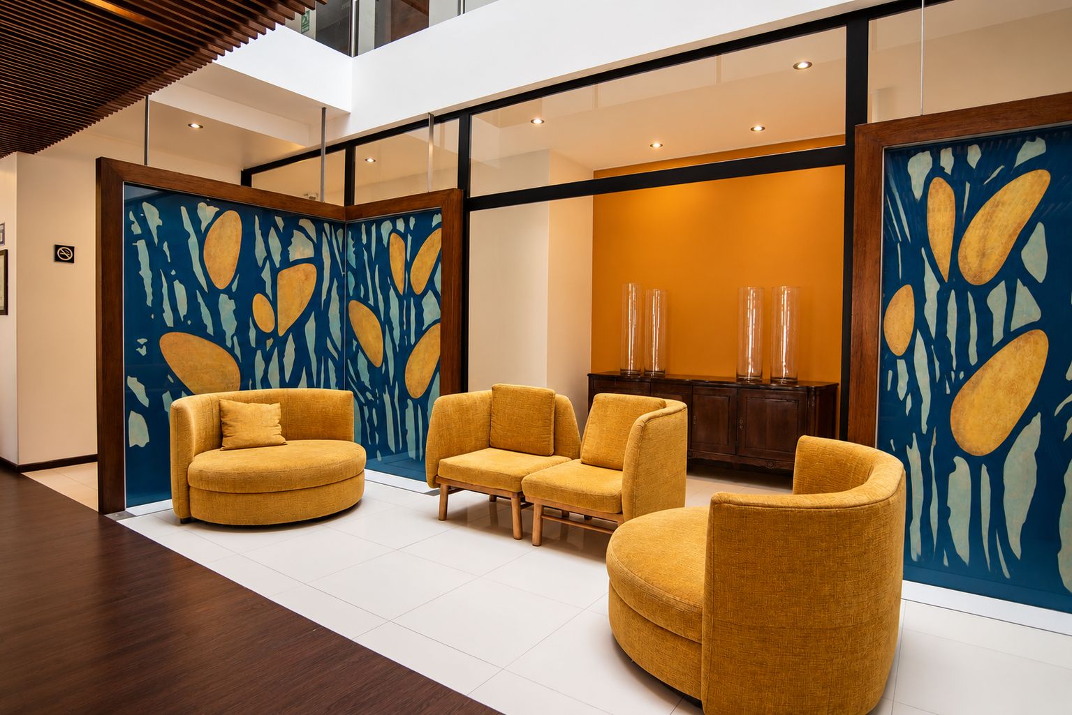 Un interior moderno con una zona de estar que incluye tres sofás y un separador de vidrio con arte abstracto.