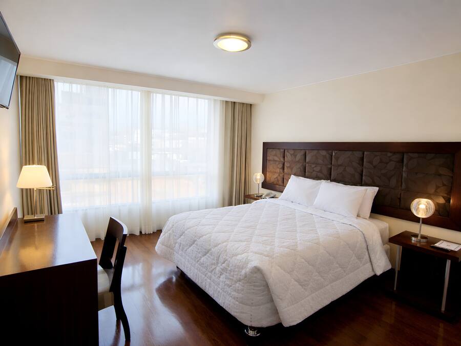 A hotel room with a bed, a desk, a chair, a TV, and a nightstand with a lamp.