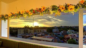 A room with a large window overlooking a cityscape at sunset, a green sofa, and a floral arrangement on the window sill.