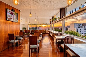 A restaurant with wooden walls, multiple tables and chairs, hanging light bulbs, and a view of the sunset through large windows. - Mint Hotel (Arequipa)