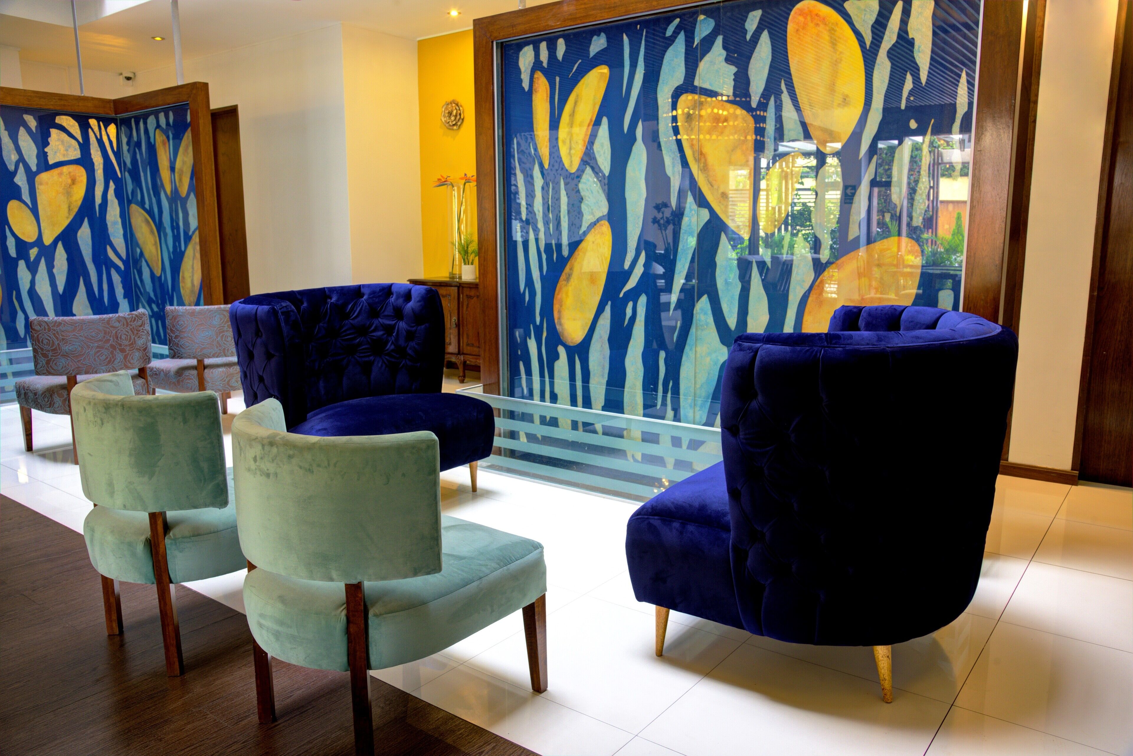 Un área de estar con sillones tapizados en azul y verde, una partición de vidrio con una obra de arte abstracta en amarillo y azul, y vista al exterior a través del vidrio.