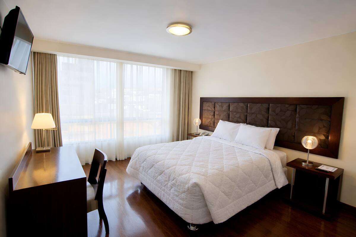 A hotel room with a bed, a desk, a chair, a TV, and a bedside lamp.