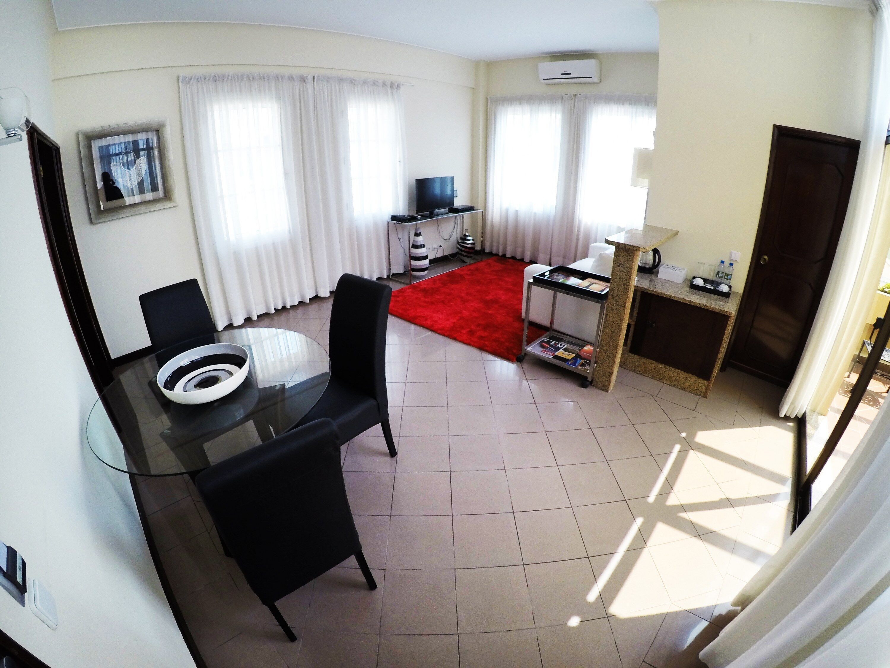 A compact hotel room with a dining area, a TV, a small kitchenette, and a red rug.