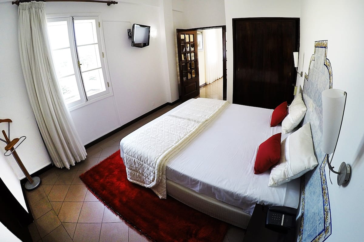 Habitación de hotel con cama, televisión, ventana con cortinas, alfombra roja y puerta que da a otra habitación.