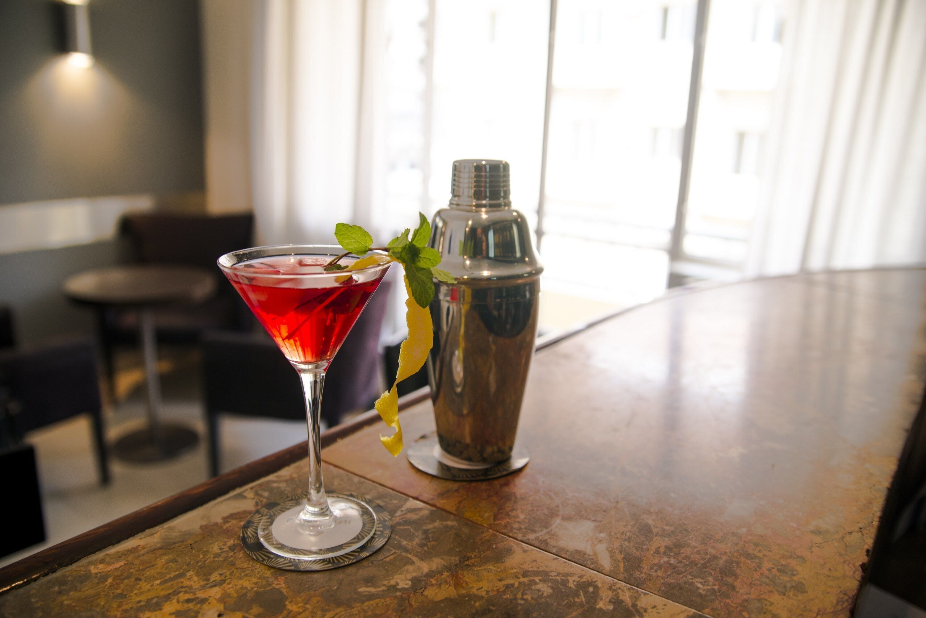 Un verre à martini contenant une boisson rouge, posé sur le comptoir d’un bar, un shaker à cocktails, et un espace bar avec des chaises et une fenêtre en arrière-plan.
